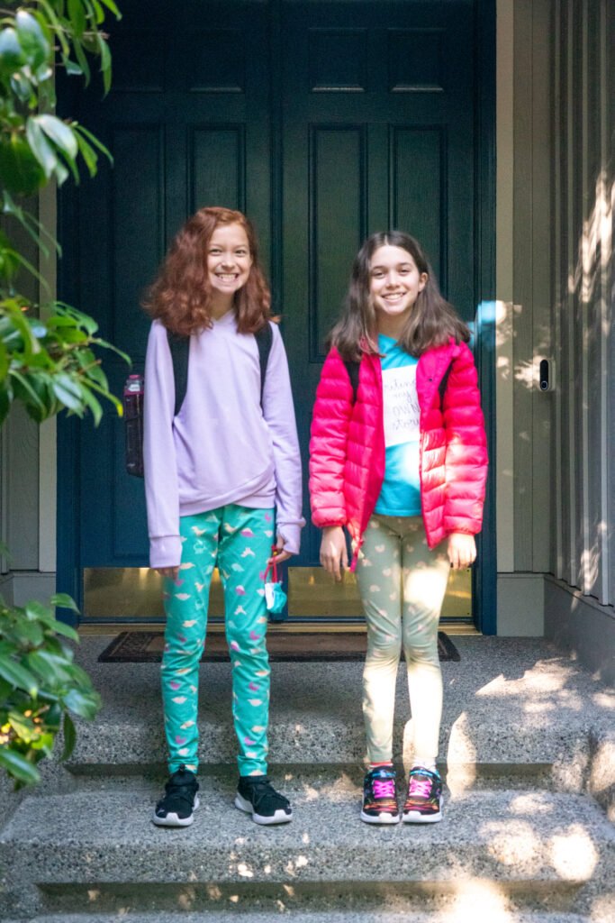 Two girls standing on the porch steps in front of a dark blue door, wearing colorful outfits and backpacks.