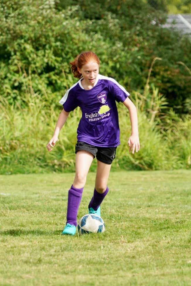 A young soccer player dribbles a ball on a grassy field while wearing a purple jersey and mismatched socks.