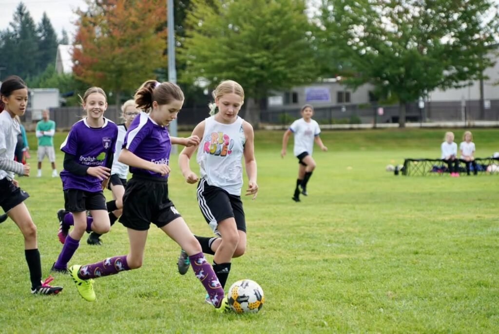 Several young players in purple and white uniforms compete for the ball on a grassy soccer field.