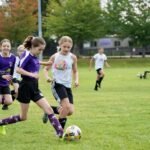 Several young players in purple and white uniforms compete for the ball on a grassy soccer field.