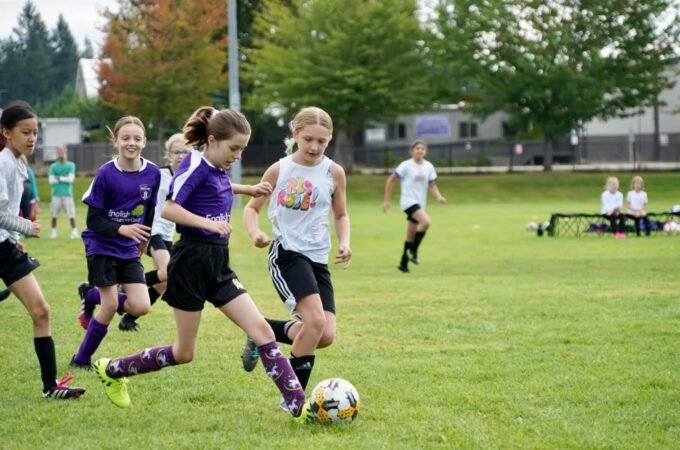 Several young players in purple and white uniforms compete for the ball on a grassy soccer field.