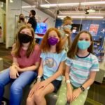 A group of three people wearing masks sits in a hospital waiting area. Nearby, others wait in a busy corridor.