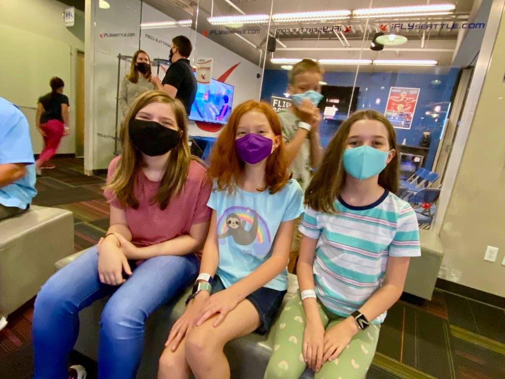 A group of three people wearing masks sits in a hospital waiting area. Nearby, others wait in a busy corridor.