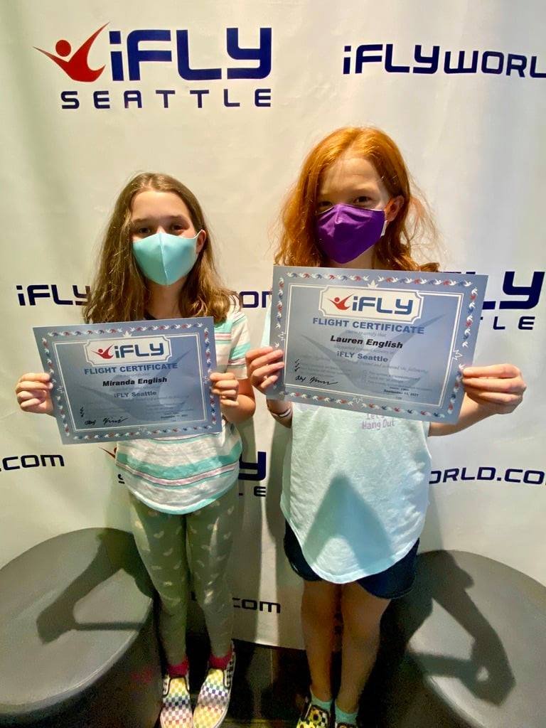 Two face-masked individuals proudly holding flight certificates and surrounded by iFLY Seattle branded backdrops and logo walls.