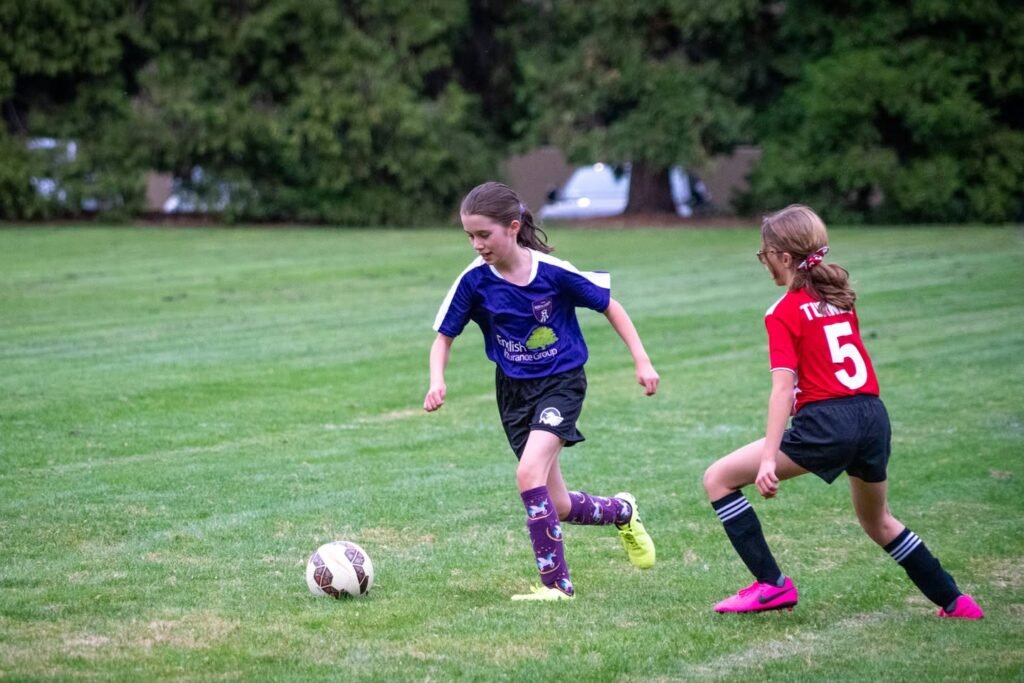 Two young players in blue and red jerseys competing for the ball on a grassy outfield. The image captures dynamic movement during a children's soccer game.