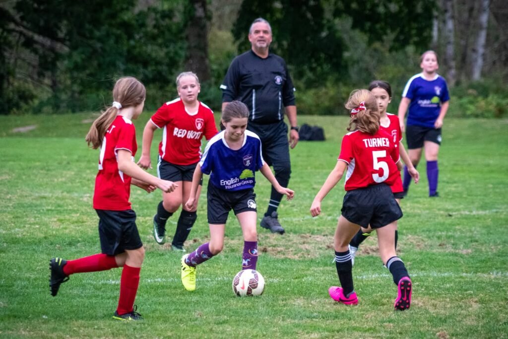 A youth soccer player dribbles the ball with opponents in red uniforms nearby, under the watchful eye of a referee in black. The players are competing on a grassy field.