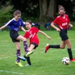 Young soccer players compete on a grassy field, with one player kicking the ball while others observe.