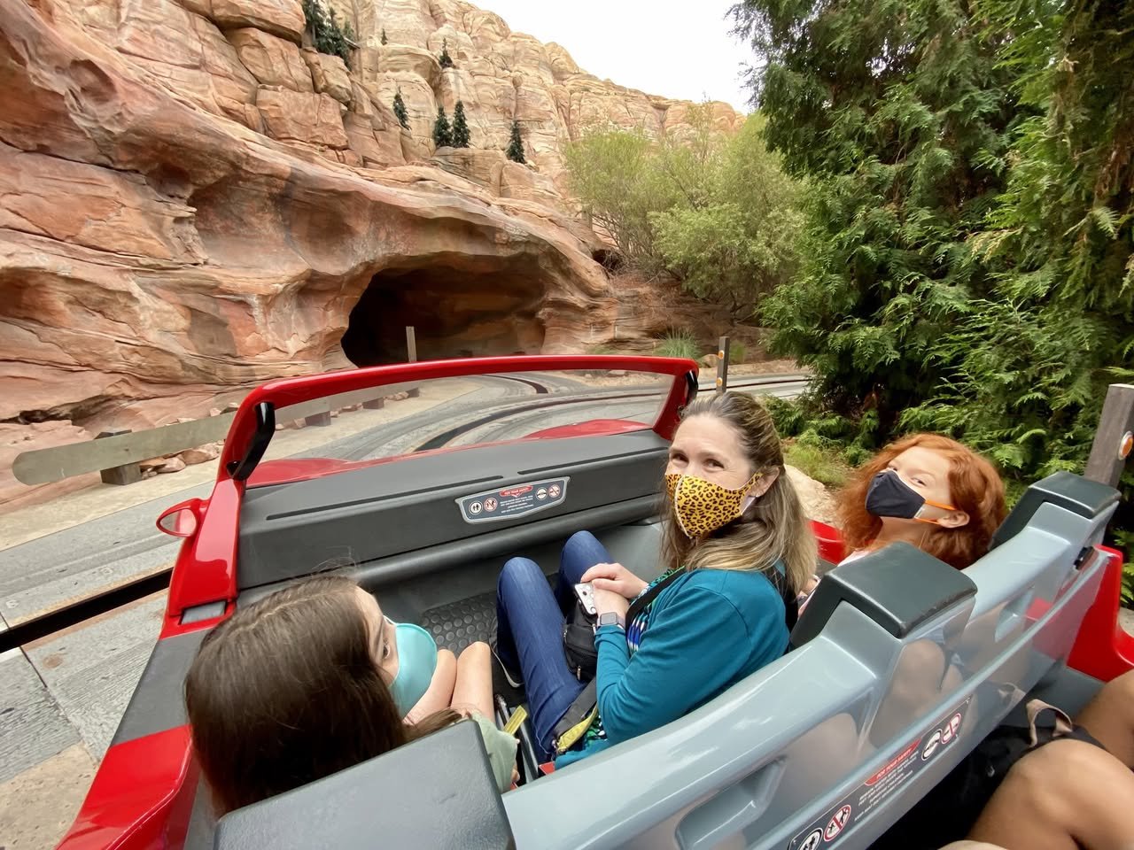 Disney California Adventure Park Family enjoying a mine cart ride through a rocky canyon at a theme park. Visitors wearing masks as they travel along a scenic rail track.