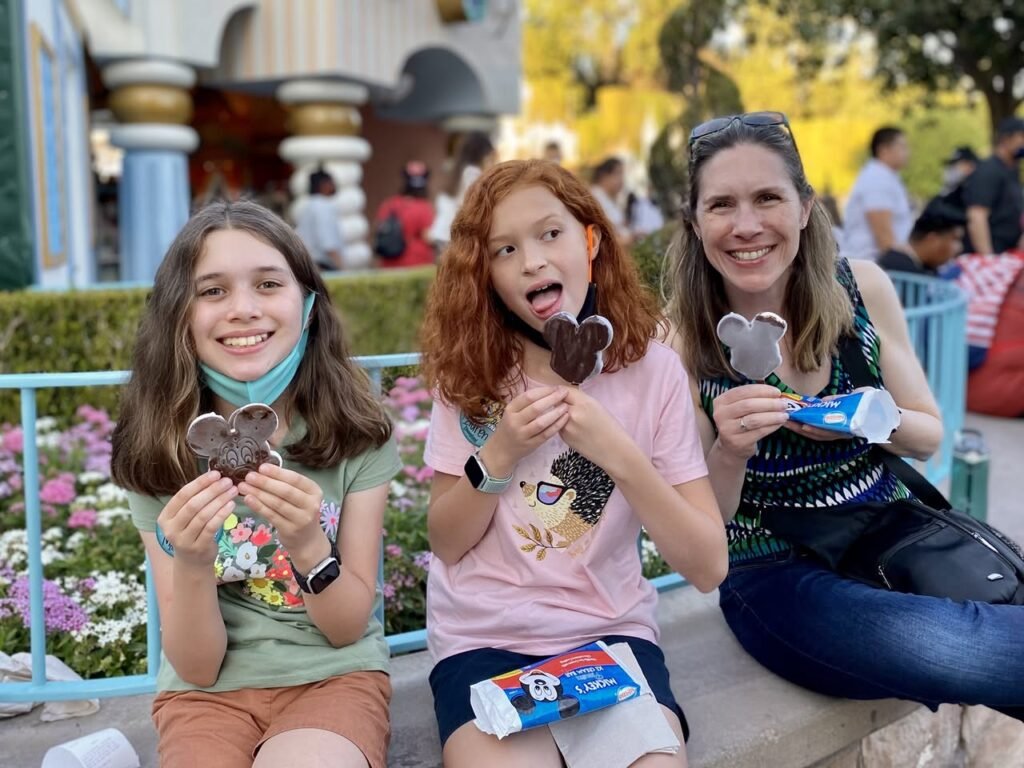 A group of three people sitting on a bench enjoying Mickey-shaped treats. The setting includes flowers and a building with columns in the background.