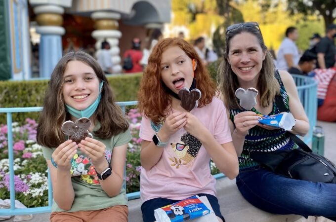 A group of three people sitting on a bench enjoying Mickey-shaped treats. The setting includes flowers and a building with columns in the background.