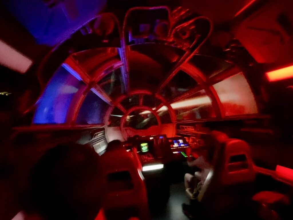A brightly lit aircraft cabin with colorful red and blue lighting. Passengers are seated in rows facing forward.