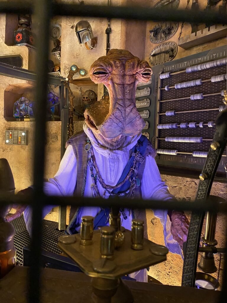 Greedo from Star Wars: A New Hope seated at a cantina table in a dimly lit Mos Eisley setting.