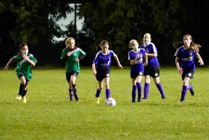 Young players in green and blue uniforms playing soccer on a well-lit field. The action shot features players chasing the ball during a competitive match.