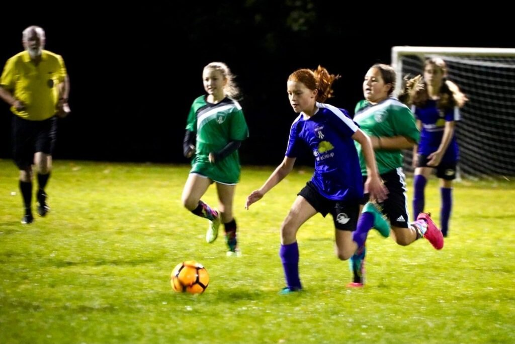 Players in green and blue jerseys compete for a ball on a floodlit field during a youth soccer match. A referee oversees the game and close-ups show players in motion.