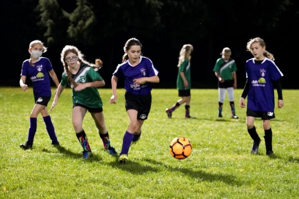 Young athletes in purple and green uniforms compete for the ball during an evening youth soccer match on a grass field.