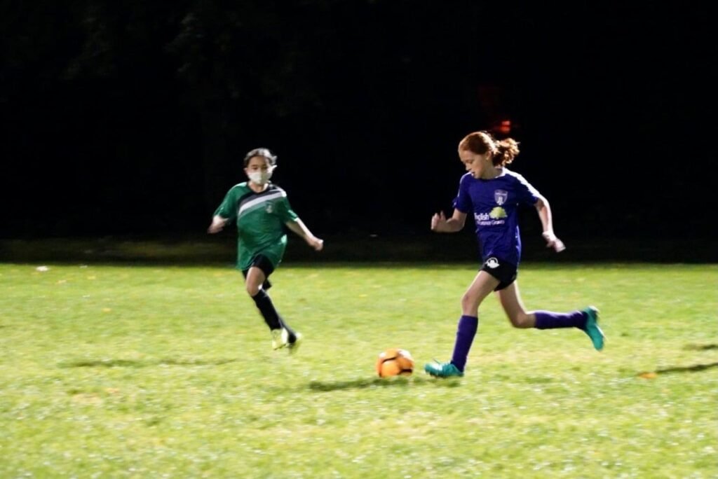 Two athletes in green and purple uniforms sprinting for an orange soccer ball.