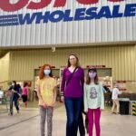 A family poses outside the Costco warehouse, with the prominent red and blue logo visible above them.