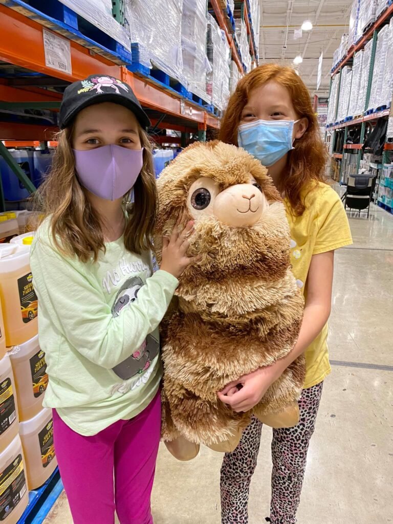 Two children wearing face masks hold a large, fluffy teddy bear in a warehouse aisle.