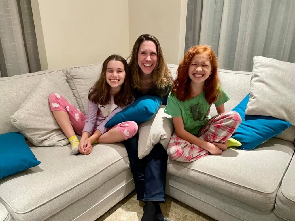 Three people relaxing together on a light beige sectional sofa with colorful pillows. Cozy indoor setting capturing a warm family moment.