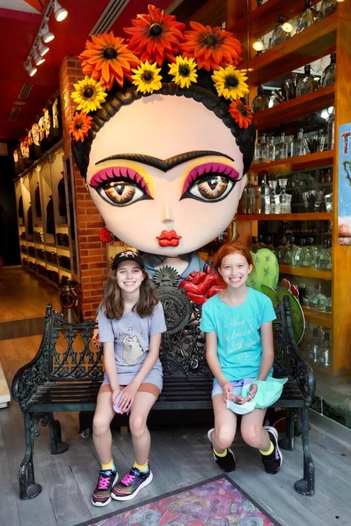 Two girls sitting on a bench in front of a large Frida Kahlo sculpture adorned with sunflowers. The setting appears to be an indoor or covered public space with brick walls and displays in the background.