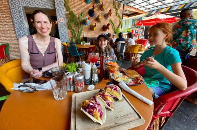 A group enjoys lunch at a colorful restaurant with tacos on a wooden tray.
