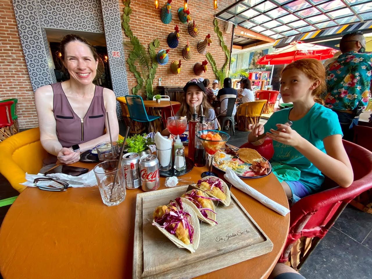 A group enjoys lunch at a colorful restaurant with tacos on a wooden tray.