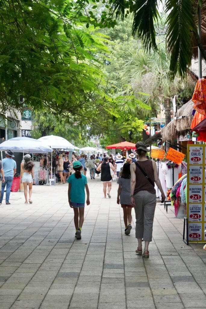 Visitors stroll through a tree-lined weekend market with colorful vendor stalls and white tents. A close-up of lush leaves adds a natural frame to the scenes.