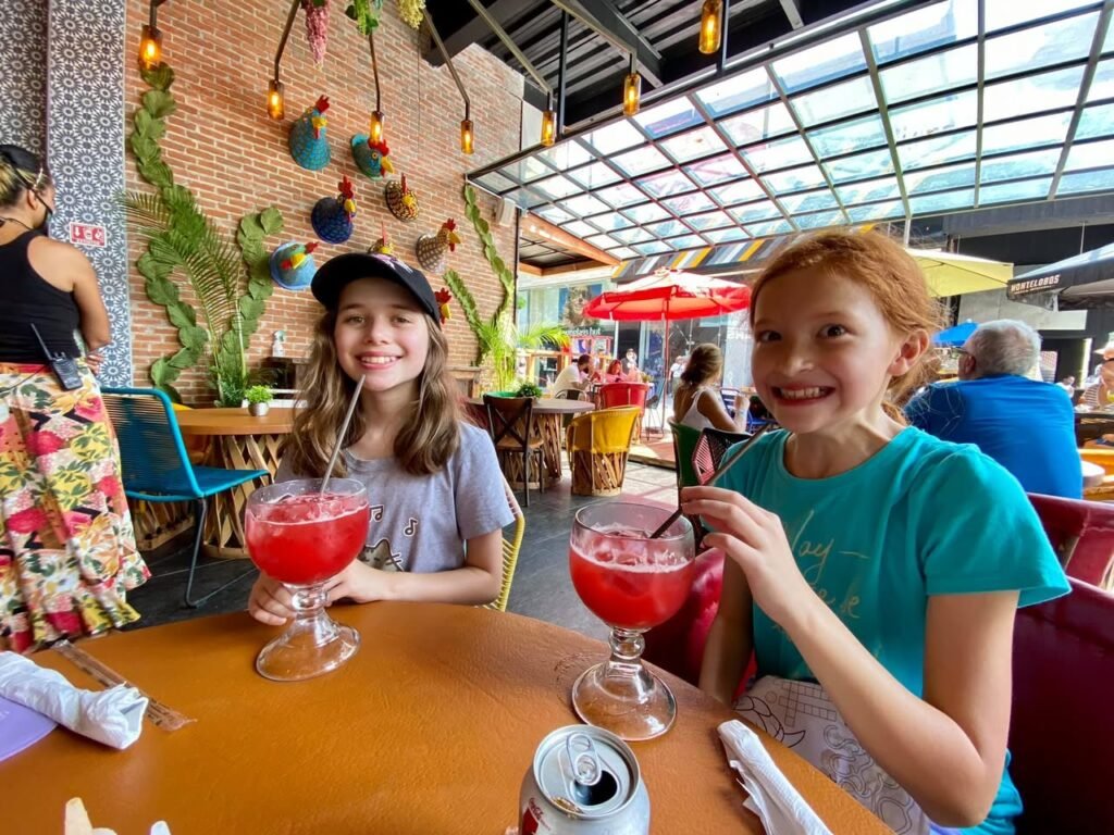 Smiling travelers enjoy red punch at a colorful eatery with a glass ceiling. The rustic decor features macrame art and tropical greenery.