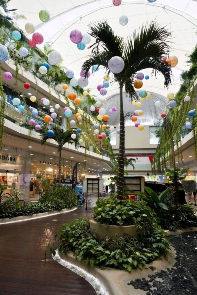 Palm trees and lush greenery decorate the interior of a bright shopping mall, with colorful hanging spheres adding a festive touch under the glass ceiling.