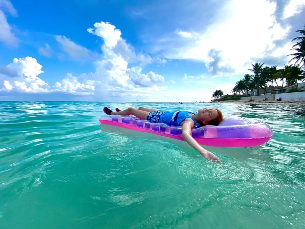 A person floats on an inflatable in clear turquoise water with palm trees nearby.