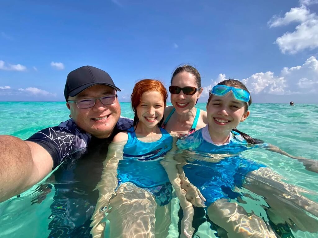 A family of four taking a selfie in the turquoise ocean water. The clear shallow sea and bright blue sky create a tropical vacation atmosphere.