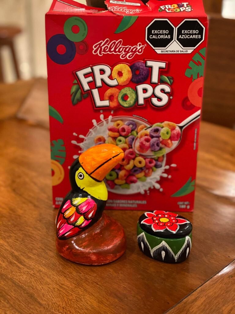 A red box of Kellogg's Froot Loops sits on a wooden table beside a vibrant toucan figurine and a painted tortoise ornament.