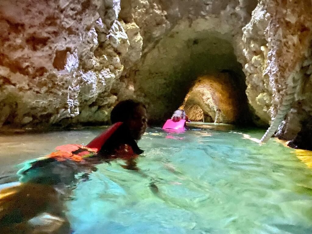 Two people kayaking through a cave with greenish water and rugged limestone walls. Textured rock formations line the cave surface, with a clear underwater view in some areas.