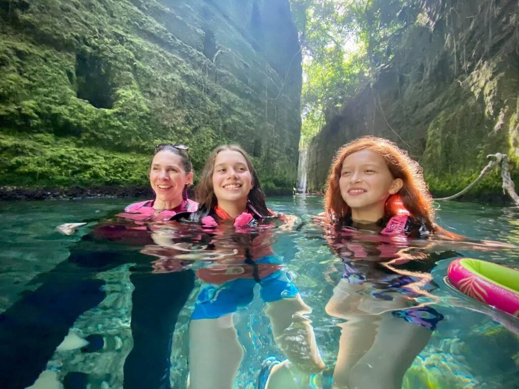 Three people wearing life vests swim in clear, turquoise water inside a lush, green grotto. A waterfall is visible in the background.
