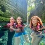 Cirque du Soleil: JOYÀ Three people wearing life vests swim in clear, turquoise water inside a lush, green grotto. A waterfall is visible in the background.
