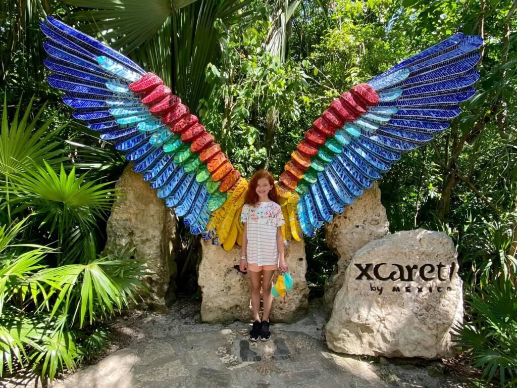 A person stands in front of large, colorful butterfly wings with a stone sign reading Xcaret by Mexcutter in a lush jungle setting.