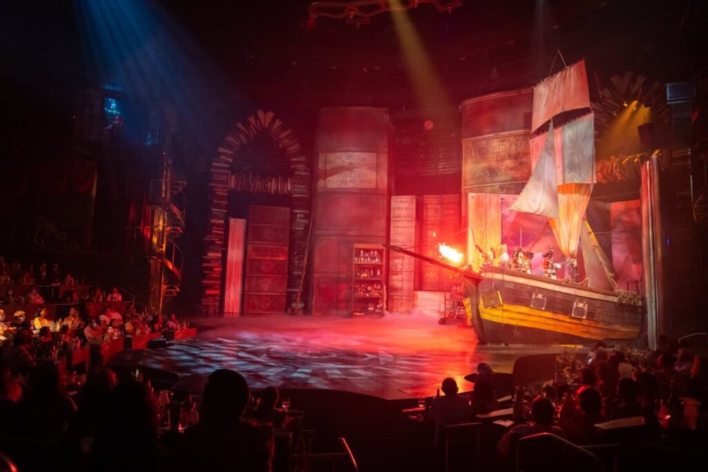 A brightly lit stage with a large ship set against a red and orange backdrop in a dark concert venue.