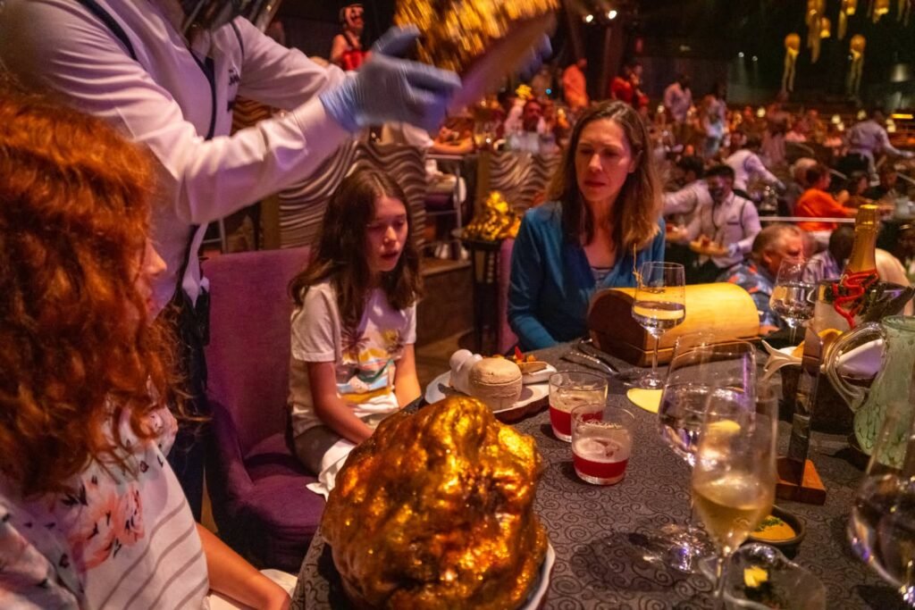A person wears a blue latex glove while cutting into a glistening golden culinary creation within a crowded, dimly lit dining room. A young girl seated nearby watches the scene unfold amid numerous other diners.