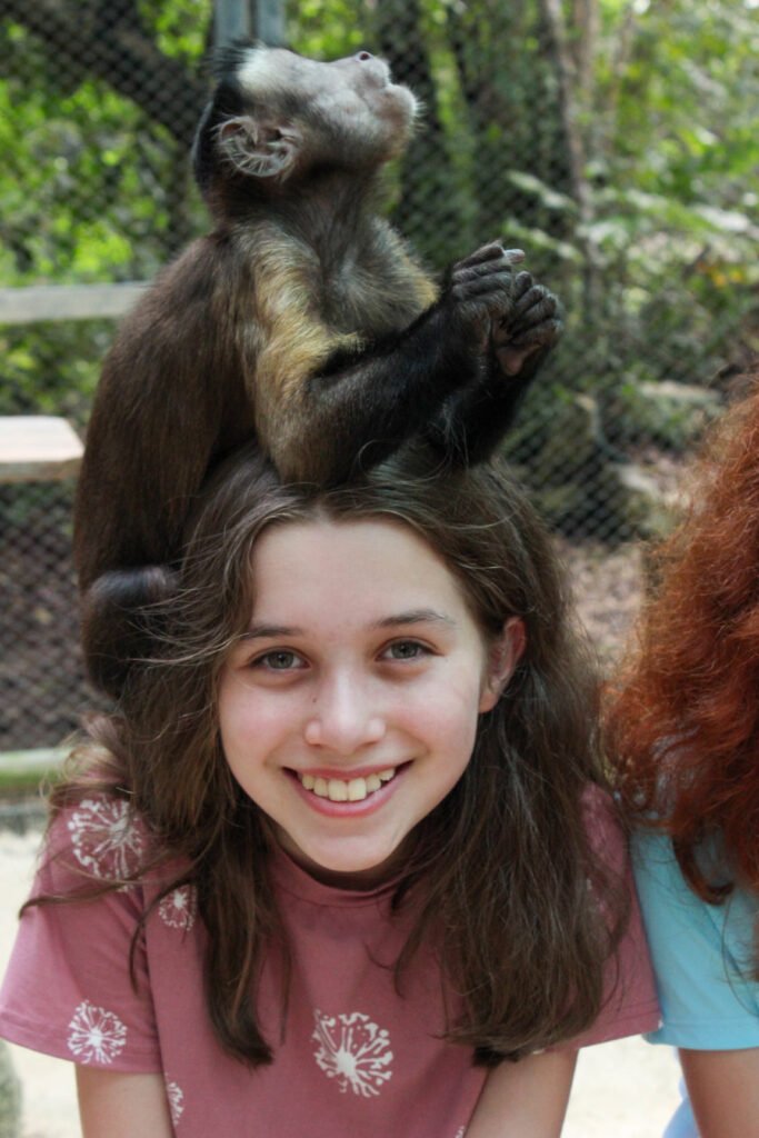 A person smiling with a small monkey perched playfully on top of their head, set against a natural outdoor background.