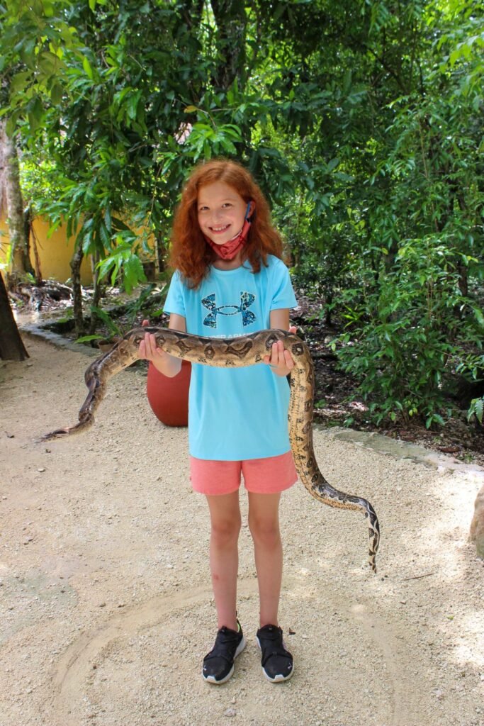 A child proudly holds a large snake at eye level while standing outdoors near greenery.