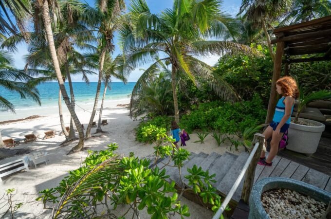 A view from a wooden deck overlooking a white sand beach with palm trees and turquoise water. A person rests on the railing near potted plants.