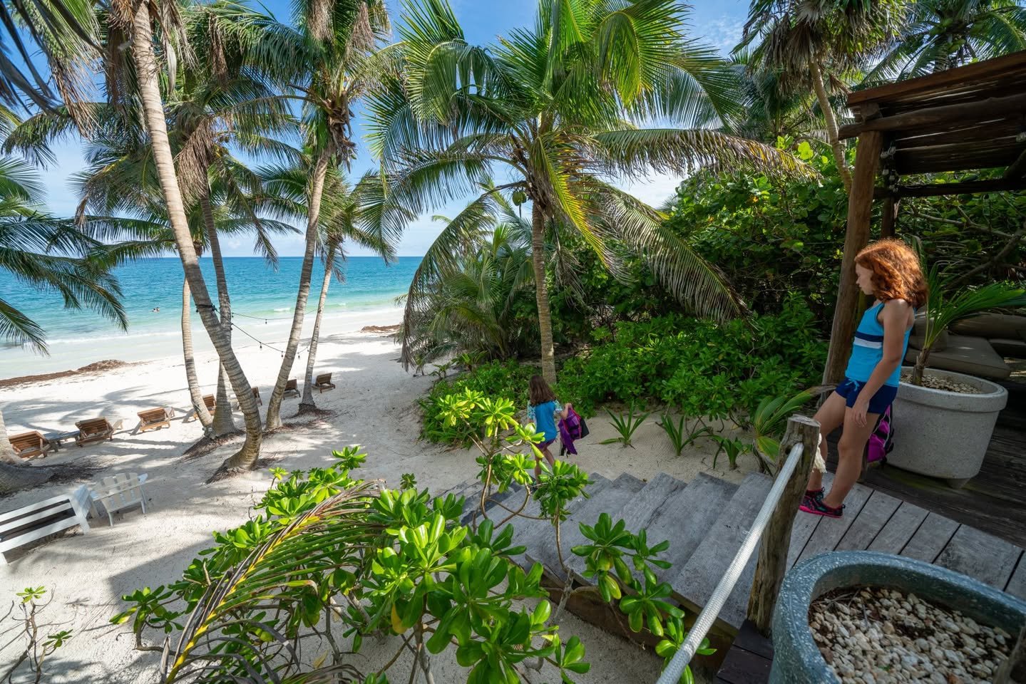 Tulum Vacation Home A view from a wooden deck overlooking a white sand beach with palm trees and turquoise water. A person rests on the railing near potted plants.