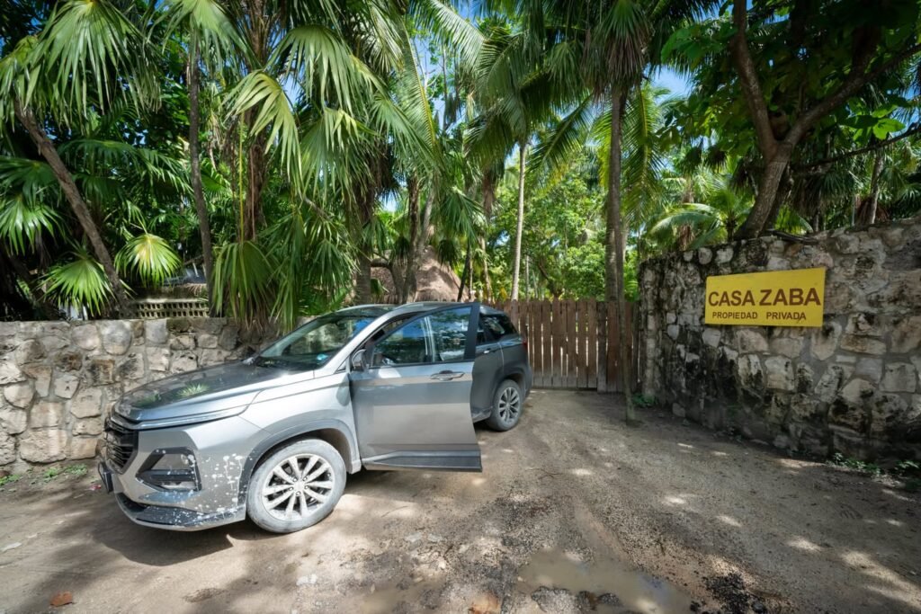 A silver SUV is parked on a dirt path next to a stone wall with a yellow sign reading 'Casa Zara'. The background features lush tropical palm trees, creating a serene entrance scene.