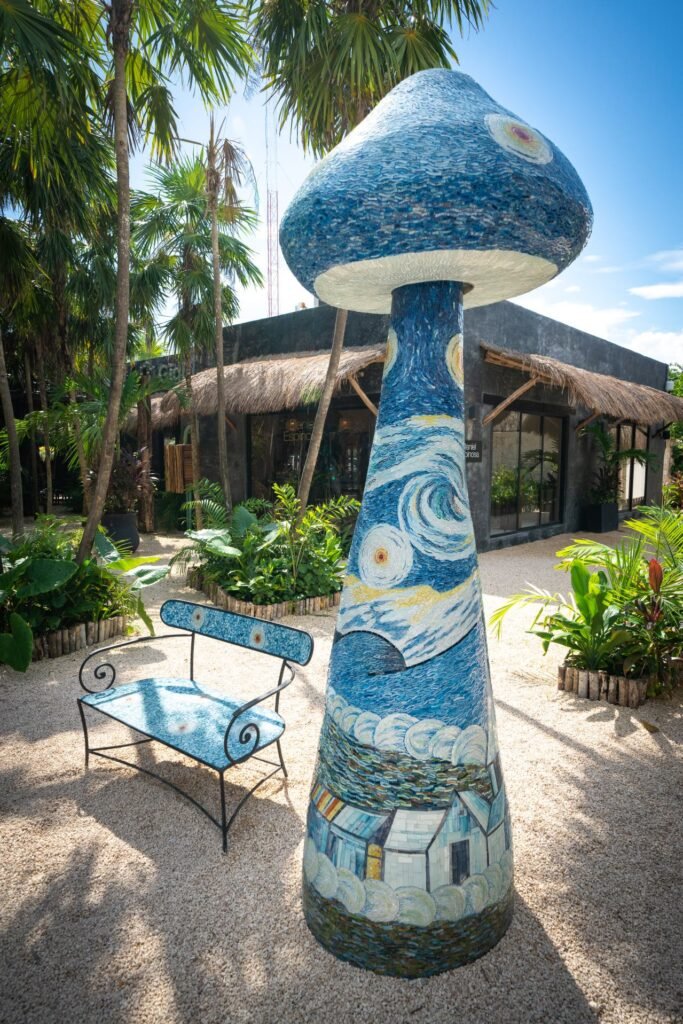 A tall, blue mosaic mushroom sculpture features artistic swirls and house motifs. Next to the sculpture is a matching ceramic bench surrounded by tropical foliage.