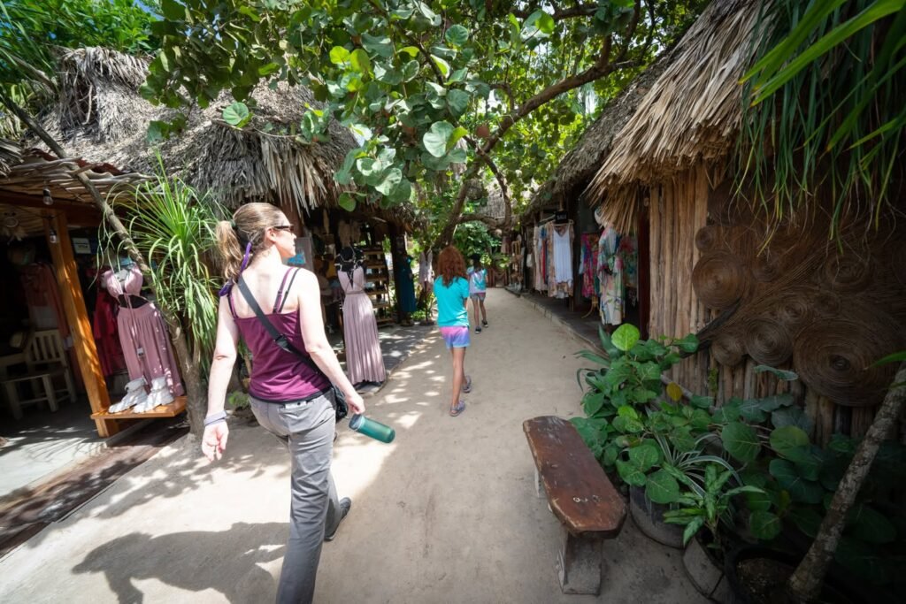 Visitors walking through a shaded tropical village with thatched-roof structures.