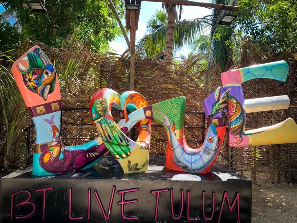 A vibrant, multicolored LOVE sign stands in a lush tropical setting with palm trees. This playful sculpture is a popular photo spot in Tulum, Mexico.