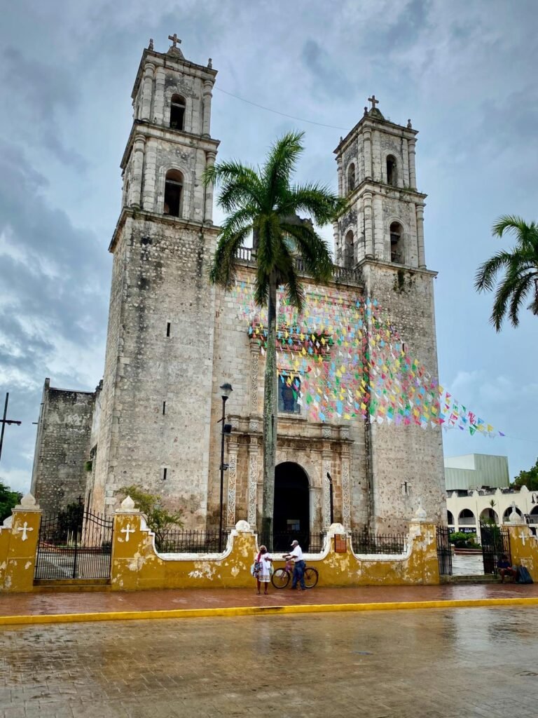 A view of the twin stone towers of the cathedral in Santiago de Cuba, featuring colorful striped banners and palm trees.