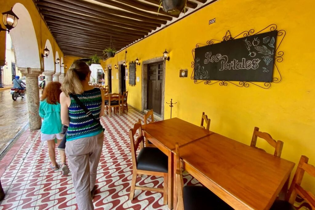 A sunny outdoor walkway with arched archways, a patterned tile floor, and a bright yellow wall featuring a chalkboard sign and wooden tables.