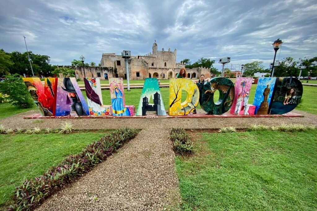 Large colorful metal sculpture spelling "Mi Amor" in a park, with the historic Mission San José building visible in the background.