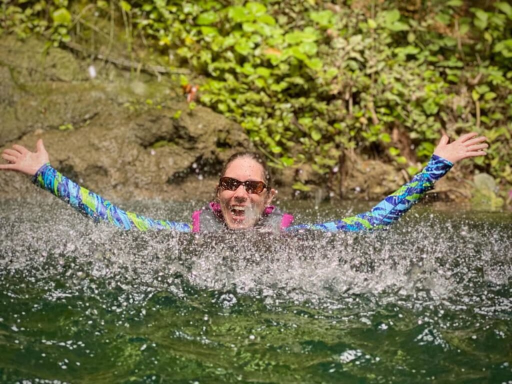 A person joyfully splashes in a clear, greenish spring with arms outstretched against a backdrop of lush foliage.